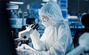 一名研究人员穿着全套防护服，戴着护目镜，在实验室的显微镜下检查样本，背景是各种科学设备。