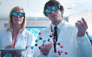 Two scientists wearing 3D glasses Two scientists, a woman and a man, stand side by side in a brightly lit, modern laboratory wearing white lab coats and large black 3D glasses. The woman, with straight blonde hair, holds a tablet in both hands and wears a red lanyard. The man has short brown curly hair and is holding a colorful molecular model made of plastic balls and sticks in black, blue, white, red, and purple, representing different atoms and bonds. He also wears a red lanyard and a name badge over a white shirt and black tie. Behind them, a digital screen displays a bright blue hexagonal molecular structure. The room is well-lit with soft white ceiling lights, pale wood walls, and a partially visible computer station in the background, giving the setting a clean and high-tech atmosphere focused on molecular research or visualization.